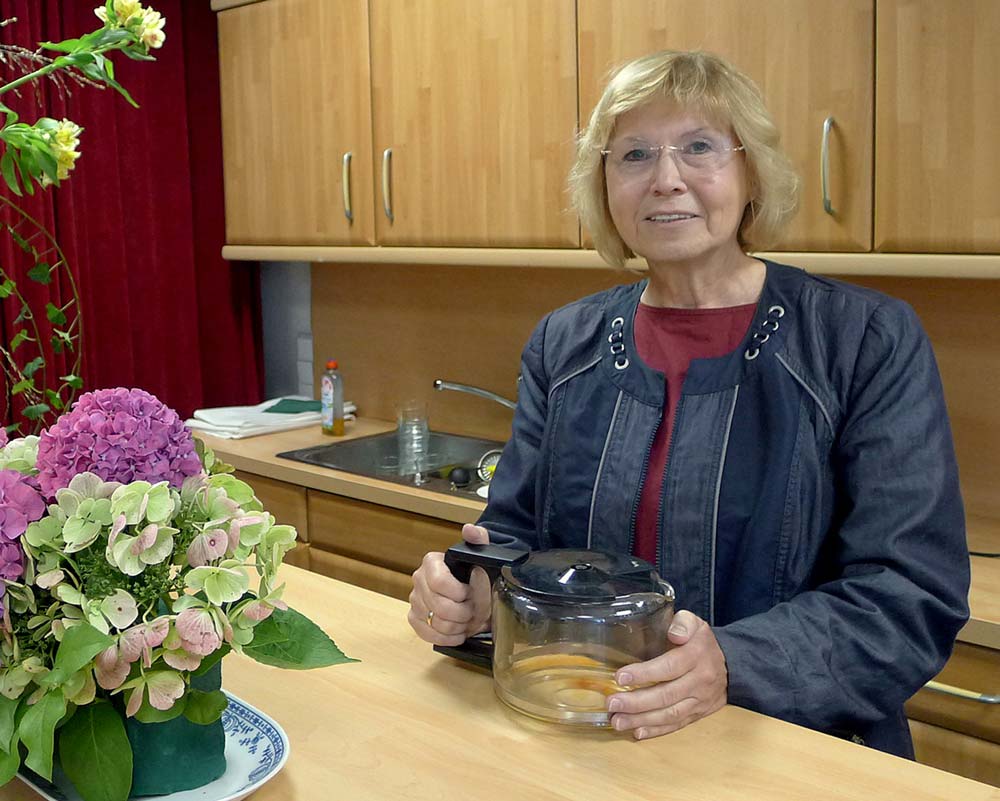 Eine Frau sitzt mit Kaffekanne und Blumen am Küchentisch.