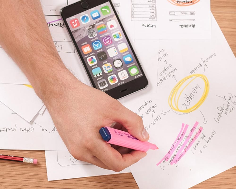 Hand mit Stift auf einer Tischplatte mit Papier und Smartphone