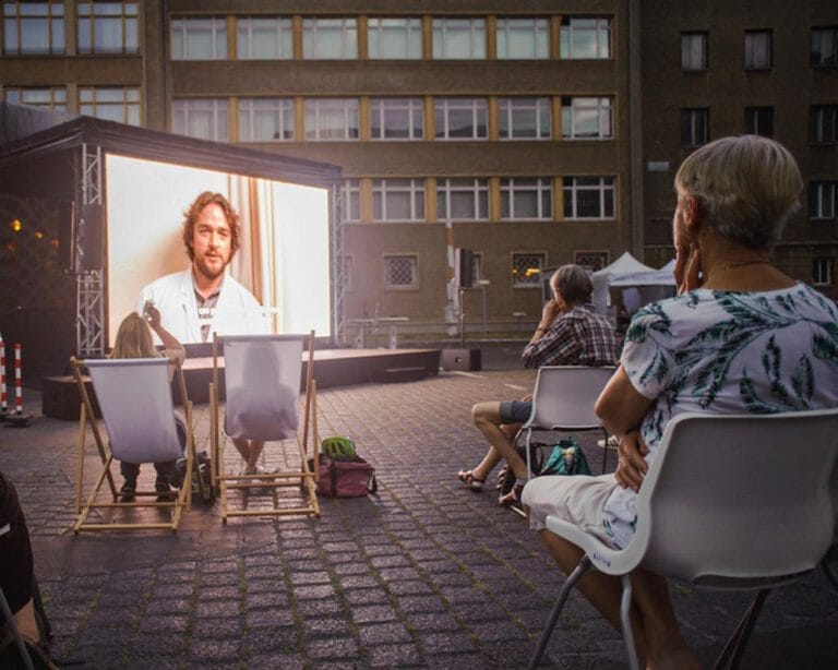 Leinwand eines Freiluftkinos, vor der menschen in Liegestühlen sitzen